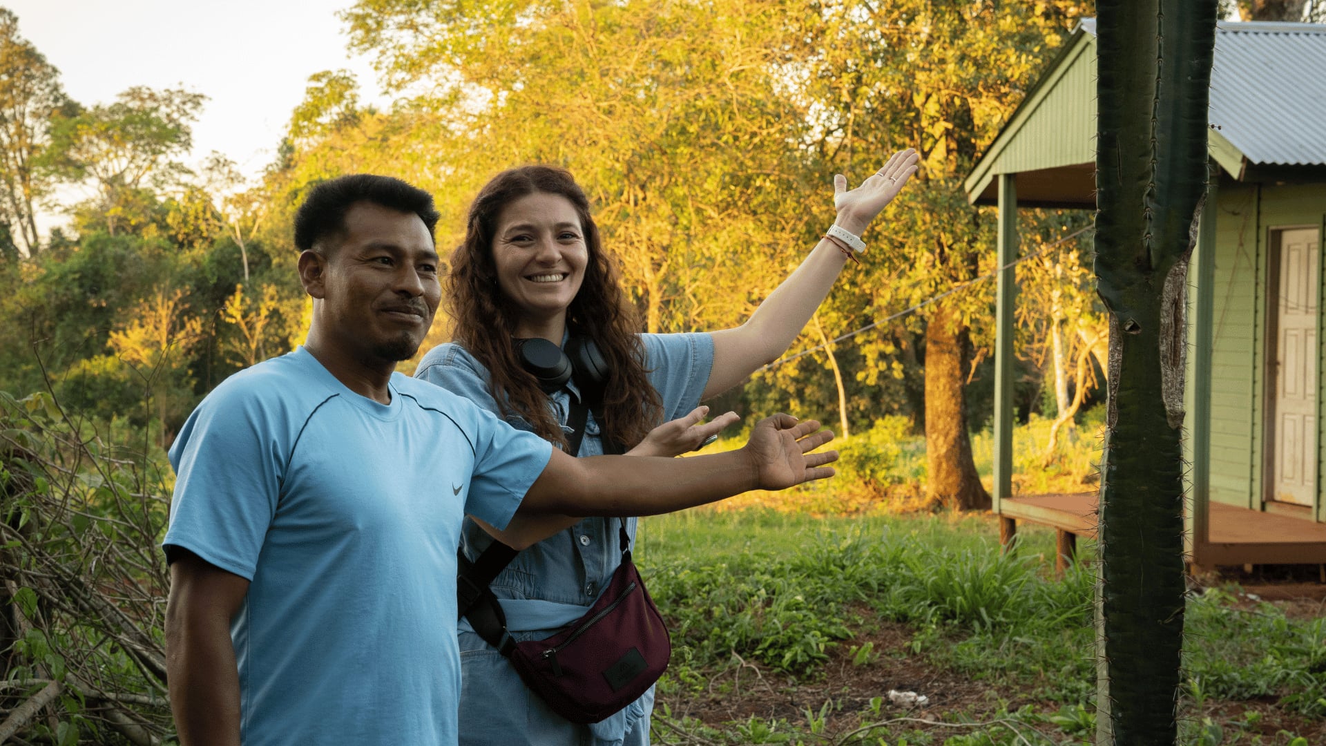 Two people smiling and gesturing toward a small house in a lush outdoor setting.