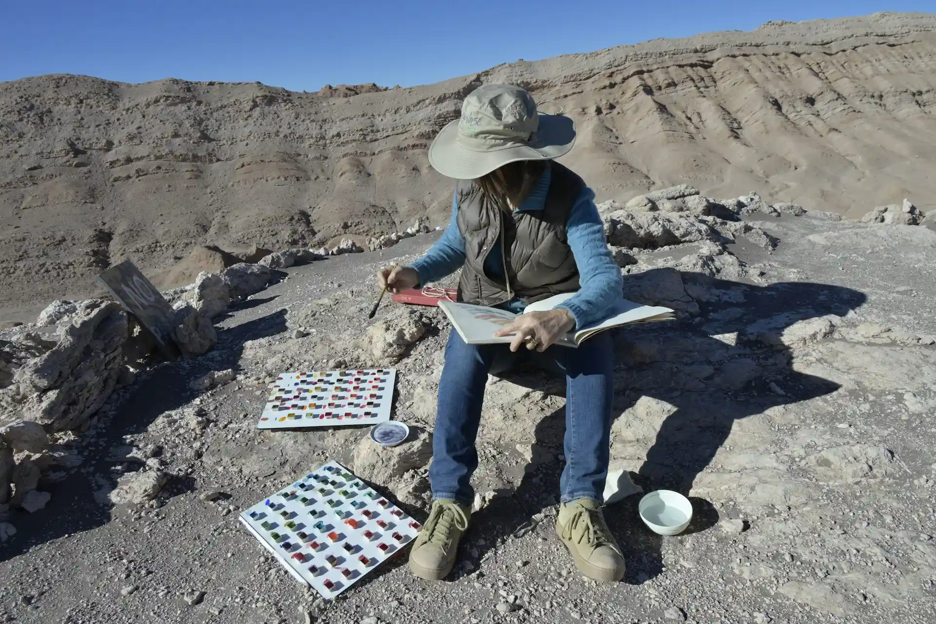 Artist seated on desert rocks studying color palettes amid layered sandstone formations.