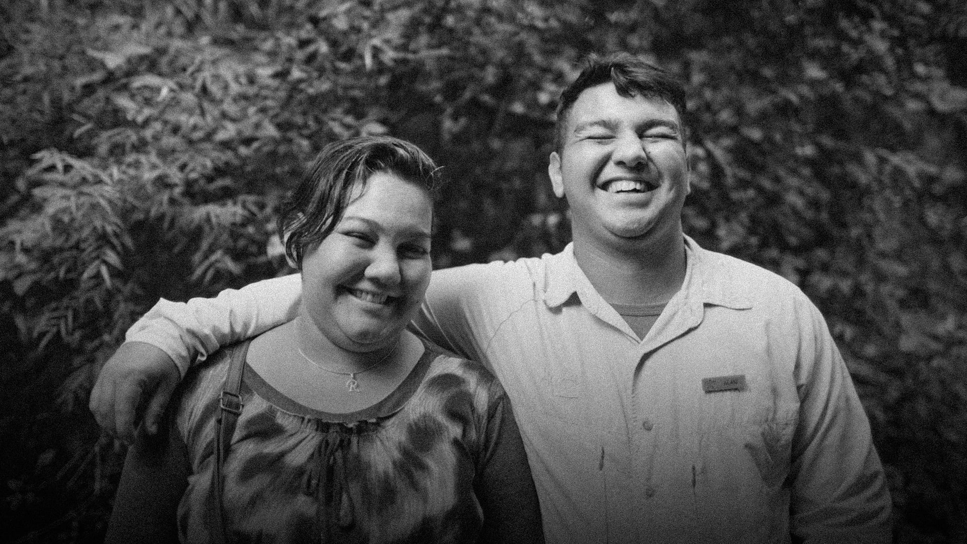 Two people smiling and laughing together outdoors, standing close with arms around each other in a natural setting.