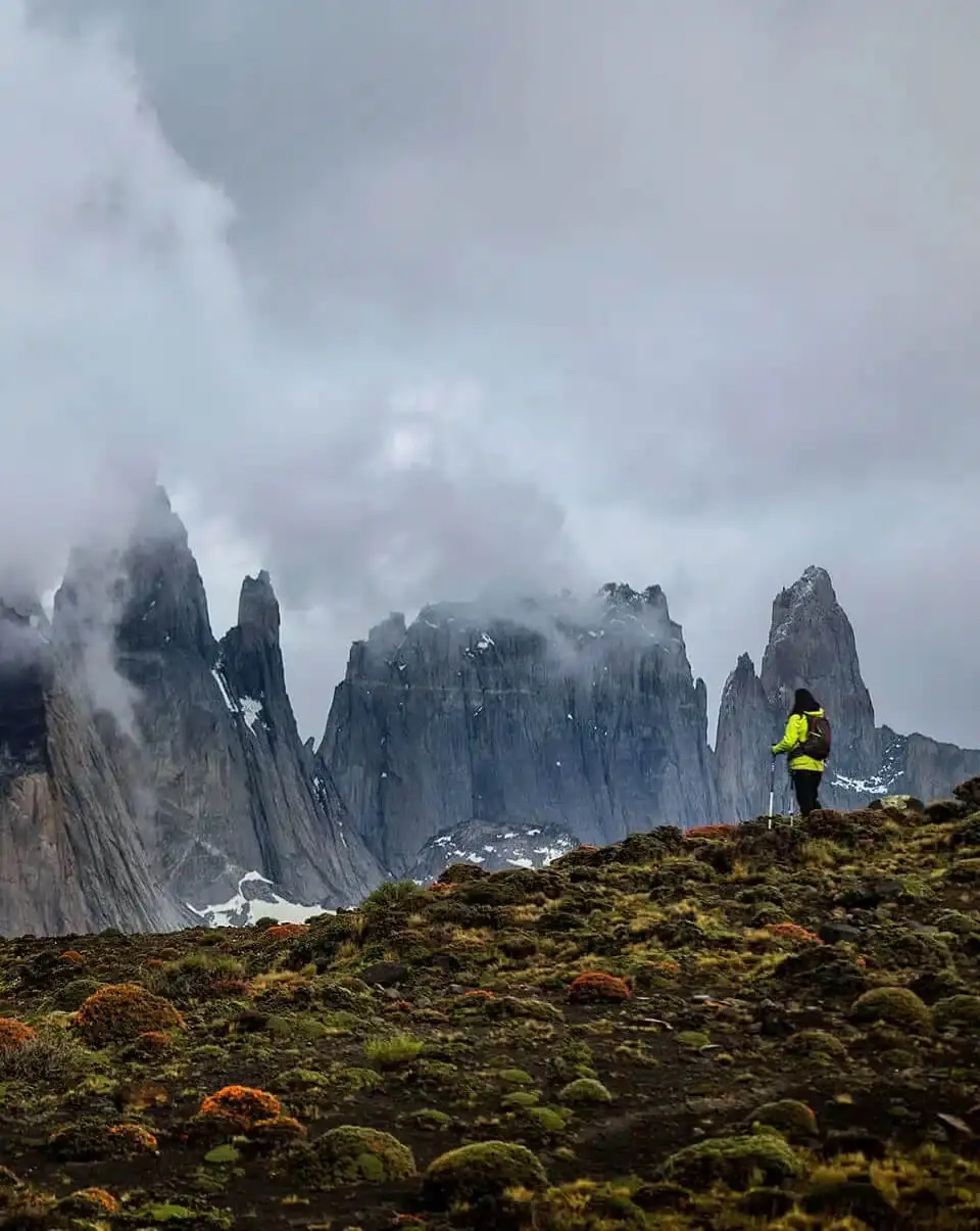 Hiker in a bright jacket standing on a mossy ridge, facing jagged mountain peaks shrouded in mist and clouds.