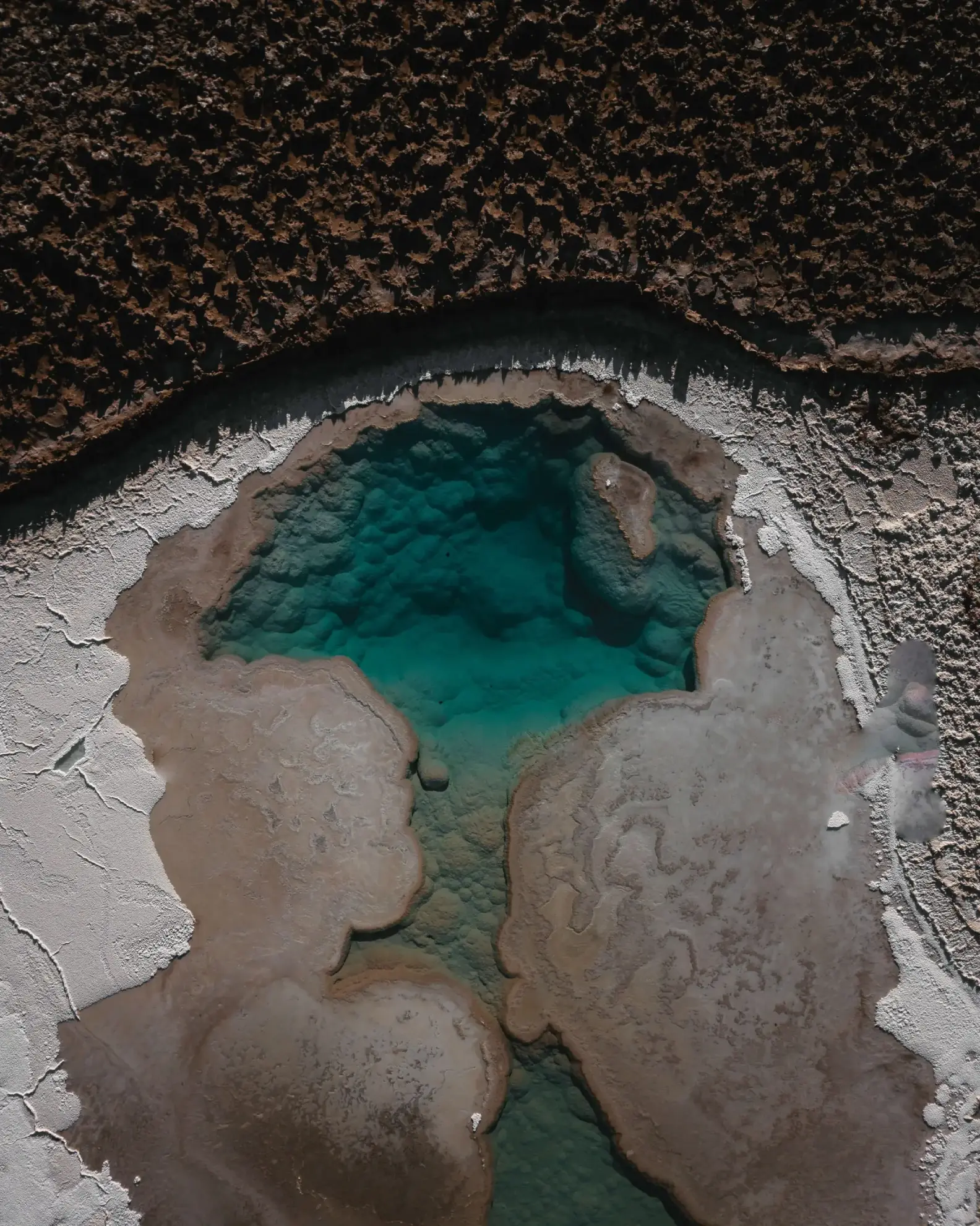 Aerial view of a turquoise mineral pool surrounded by white salt crust and dark desert terrain.
