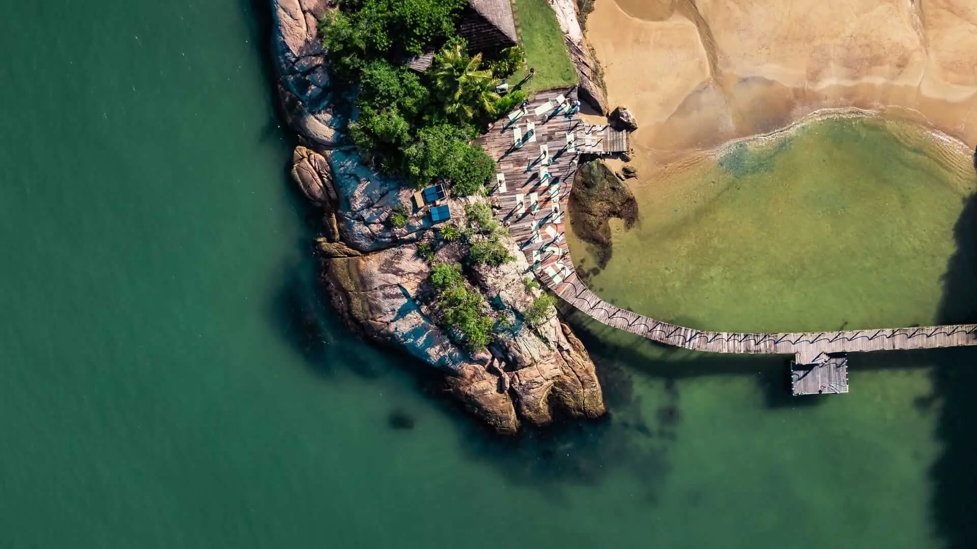 Aerial view of a rocky peninsula with wooden deck and curved pier beside shallow green water and sandy beach.