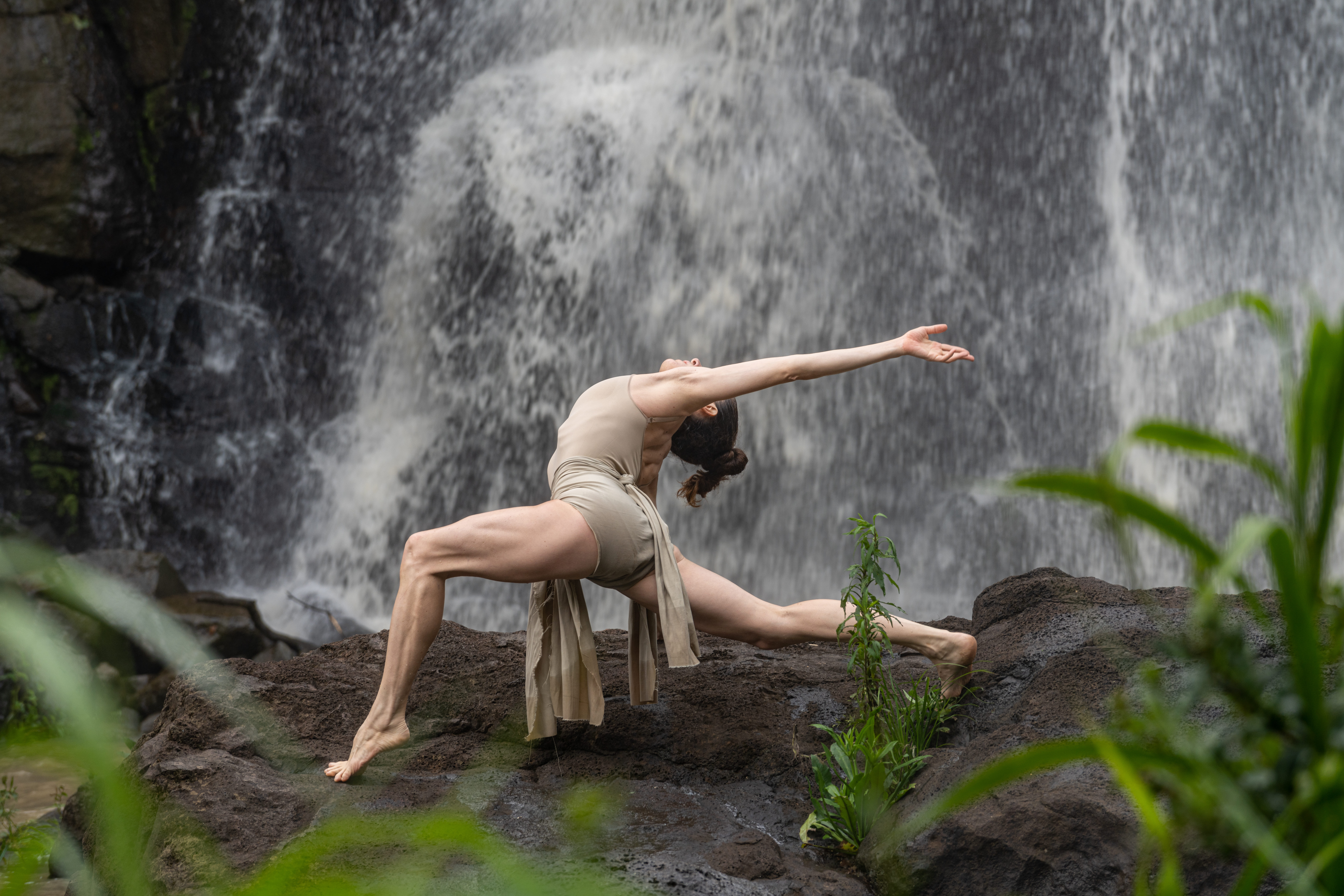 Dancer performs a graceful yoga pose on rocks beside a waterfall, surrounded by lush greenery.