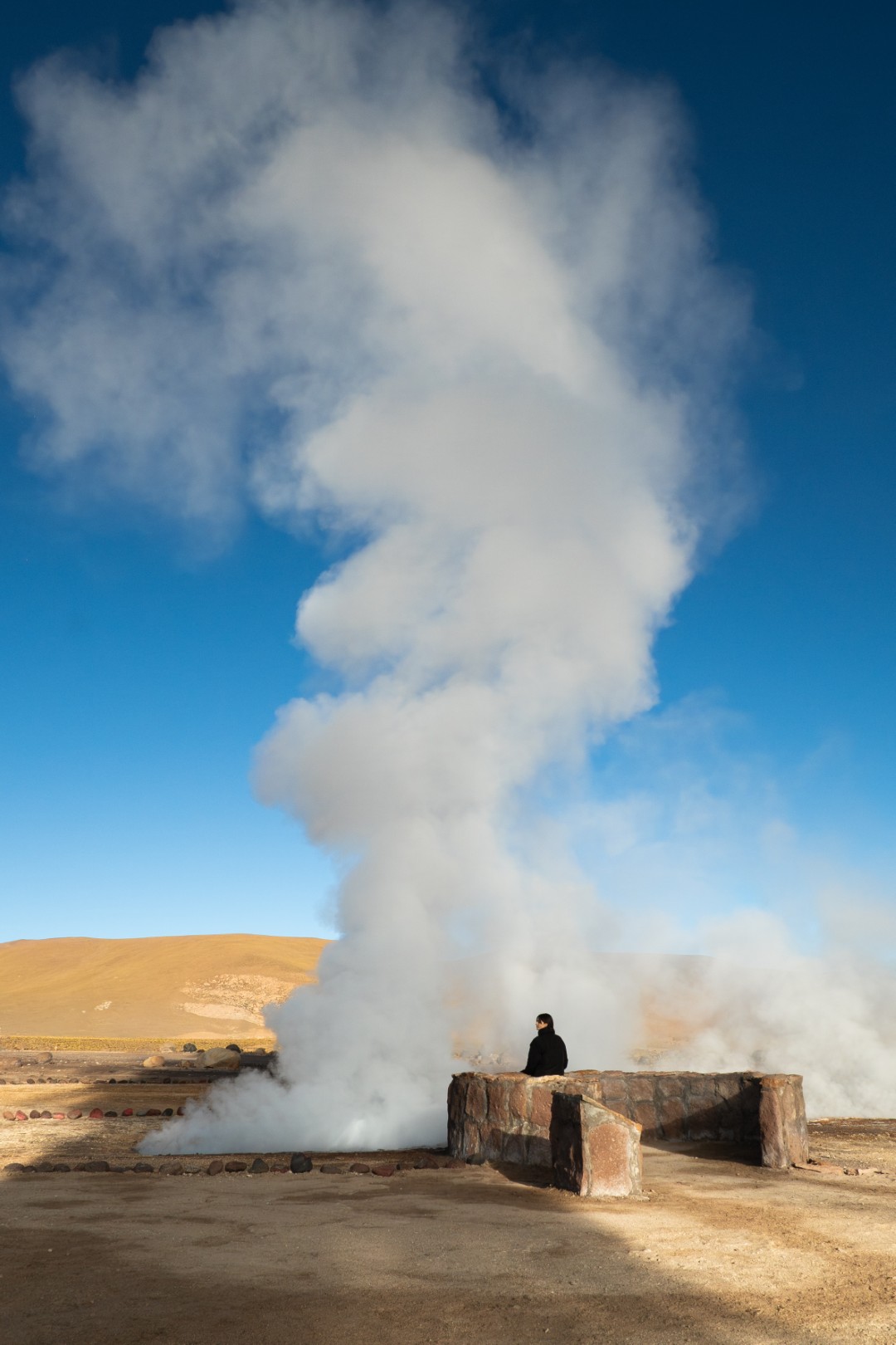 Person standing near a steaming geothermal vent in a barren desert landscape under a clear blue sky.