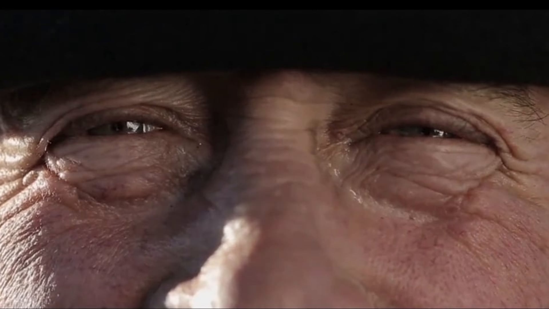 Close-up of a weathered face focusing on tired eyes beneath a dark hat, showing deep wrinkles and textured skin.
