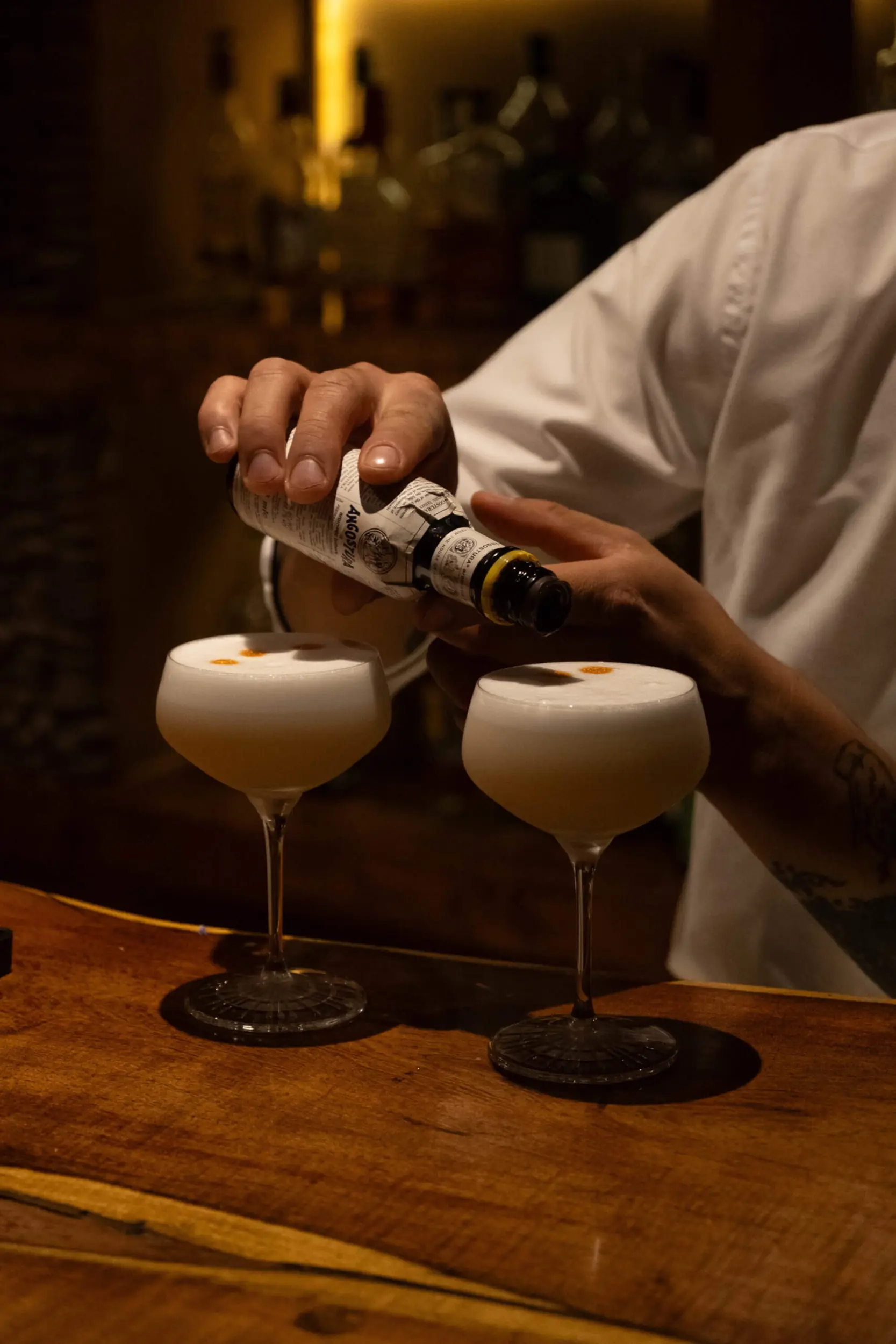 Bartender Adding Bitters To Two Foamy Cocktails In Coupe Glasses On A Wooden Bar.