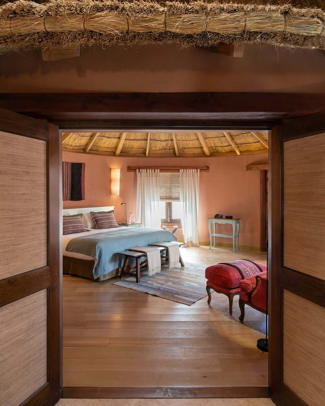 Cozy bedroom with a thatched ceiling, wooden floors, neatly made bed, and warm earthy decor seen through open doors.