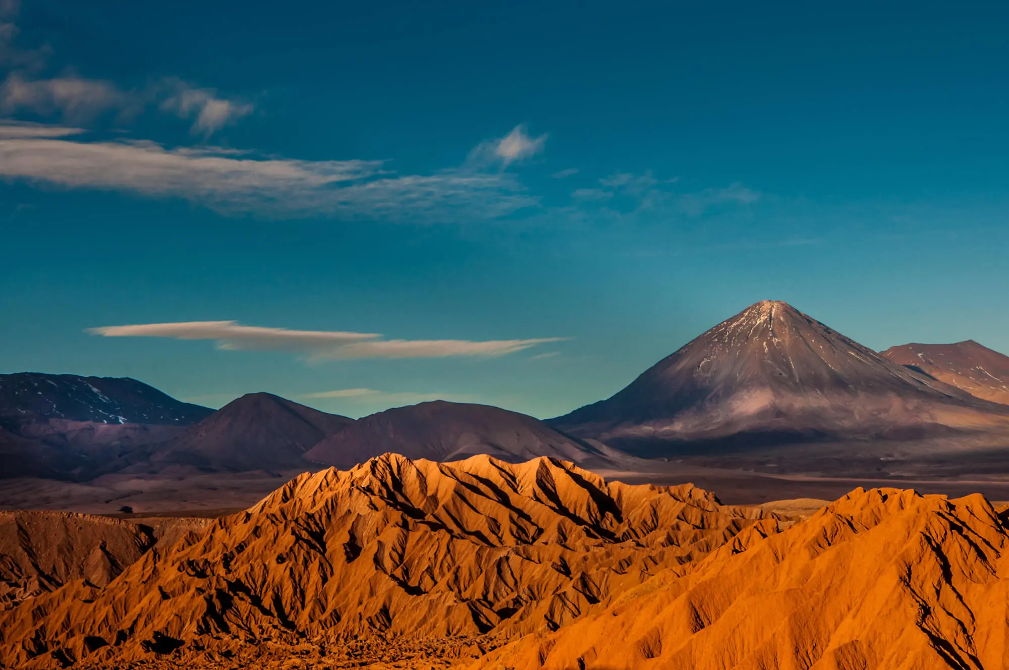 Volcanic Mountain Rising Above Golden Desert Ridges Under A Clear Blue Sky With Soft Clouds.