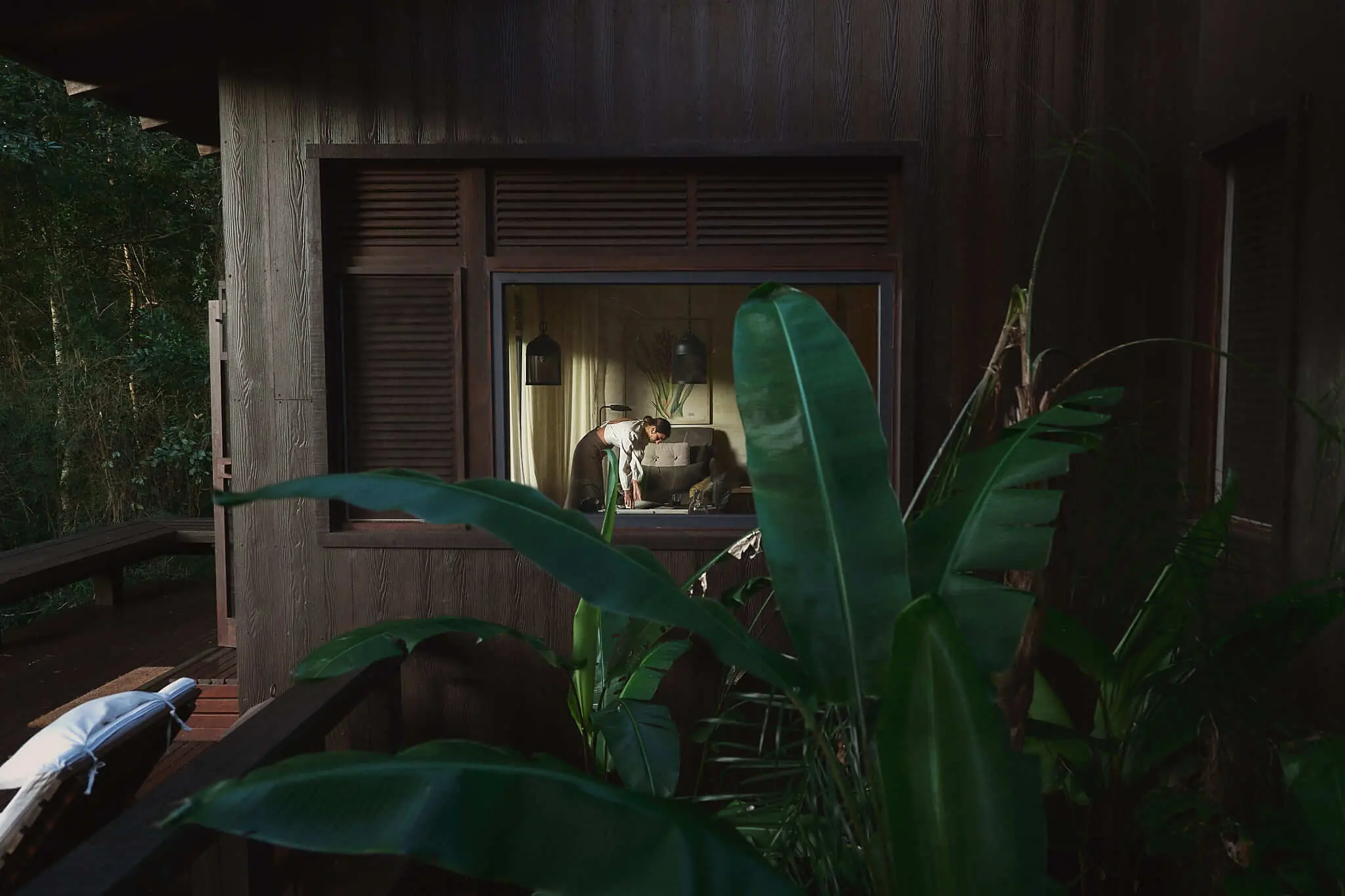 View into a jungle lodge room through a large window, with a guest preparing the space amid lush tropical plants.