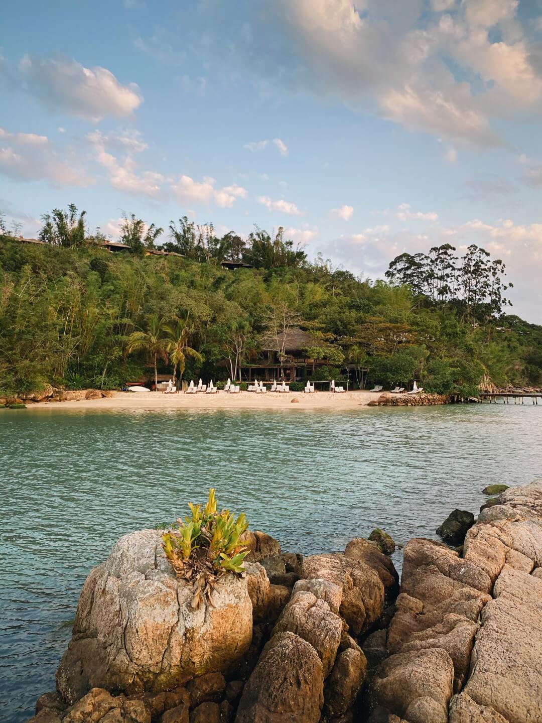 Secluded tropical beach with lounge chairs, calm water, rocky shoreline, and lush green hillside under a soft evening sky.