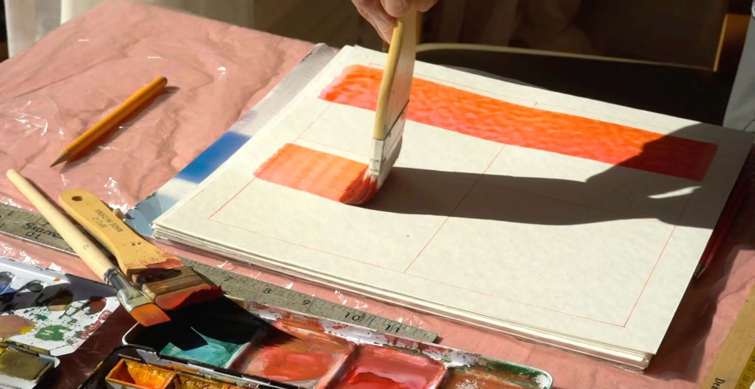 Hand painting orange watercolor swatches on gridded paper beside brushes, ruler, pencil, and paint palette.