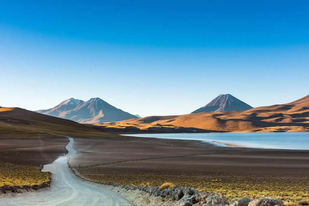 Winding Dirt Road Through A Vast Desert Landscape, With Mountains And A Calm Blue Lake Under A Clear Sky.