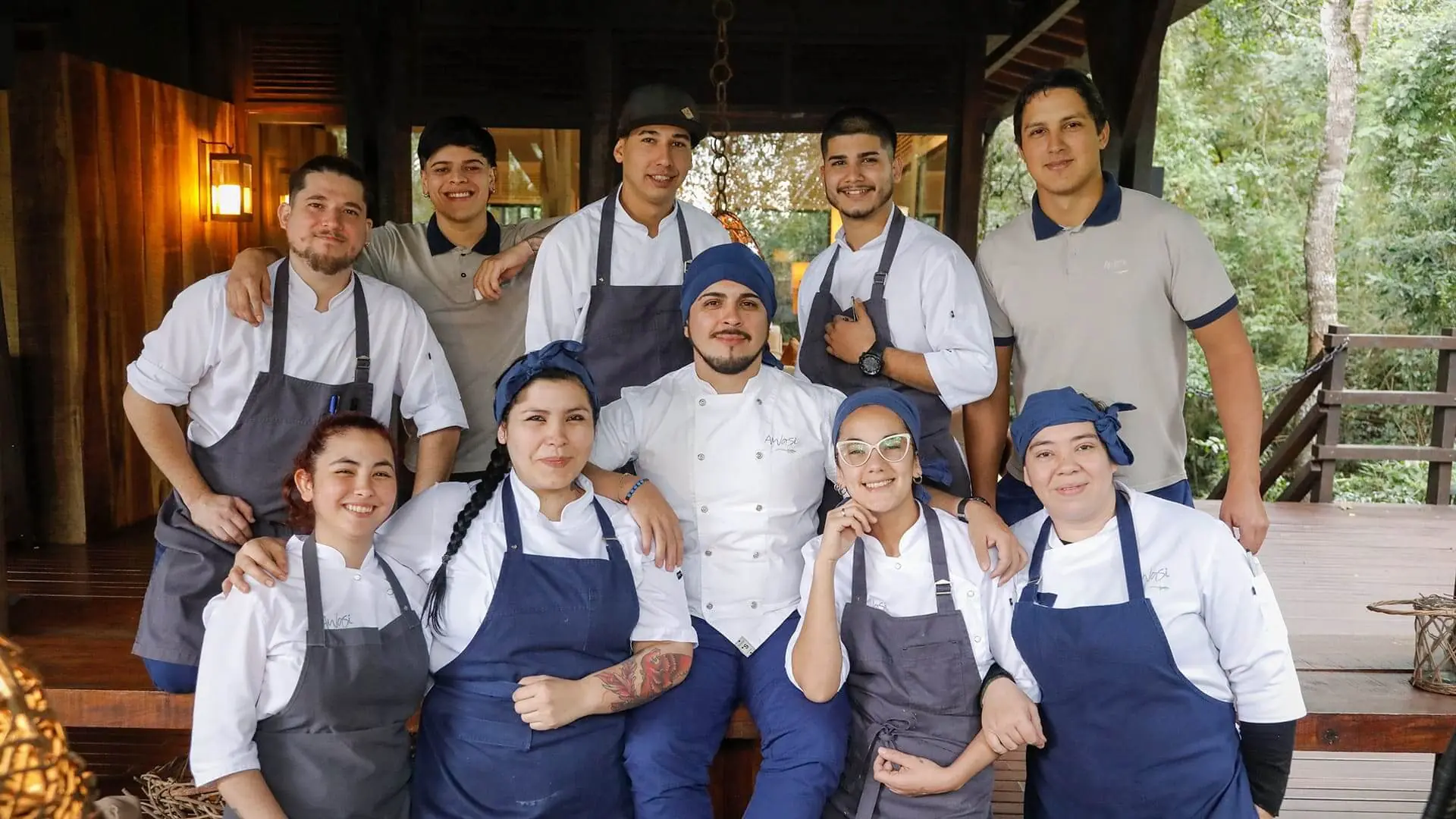 Smiling Restaurant Team Of Chefs In Uniforms Posing Together On A Wooden Terrace Surrounded By Greenery.