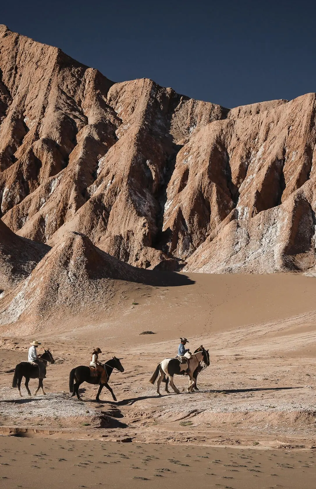Three riders on horseback crossing sandy desert beneath towering rugged rock formations.
