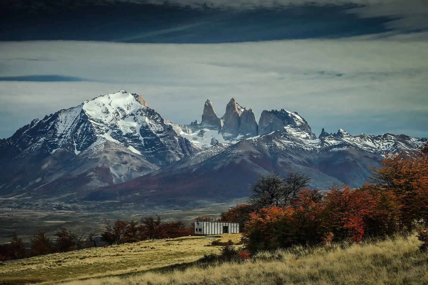 Mountain lodge set against dramatic snow-capped peaks, autumn trees, and open grassland under a moody sky.