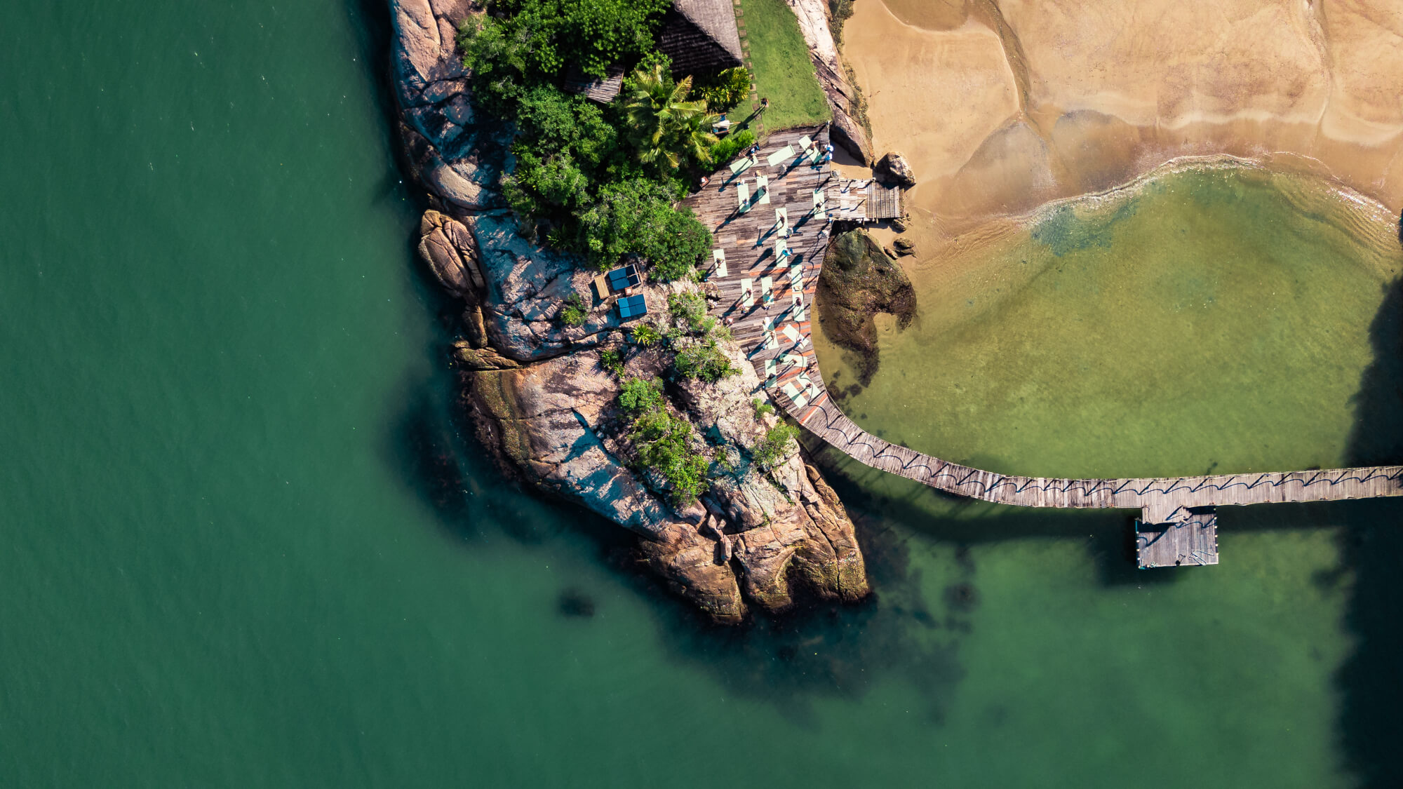 Aerial view of wooden pier, rocky shoreline, and turquoise water beside sandy beach.