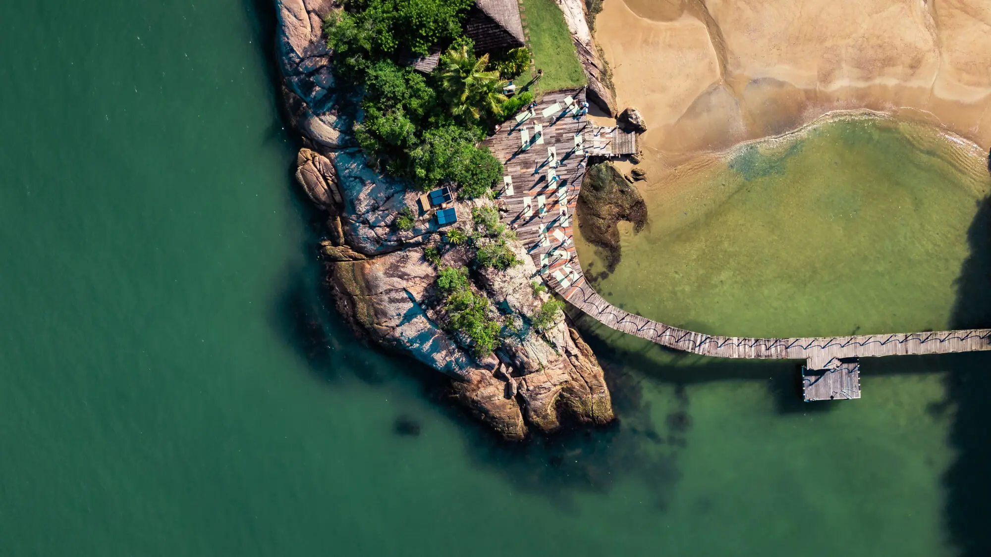 Aerial View Of Wooden Pier, Rocky Shoreline, And Turquoise Water Beside Sandy Beach.