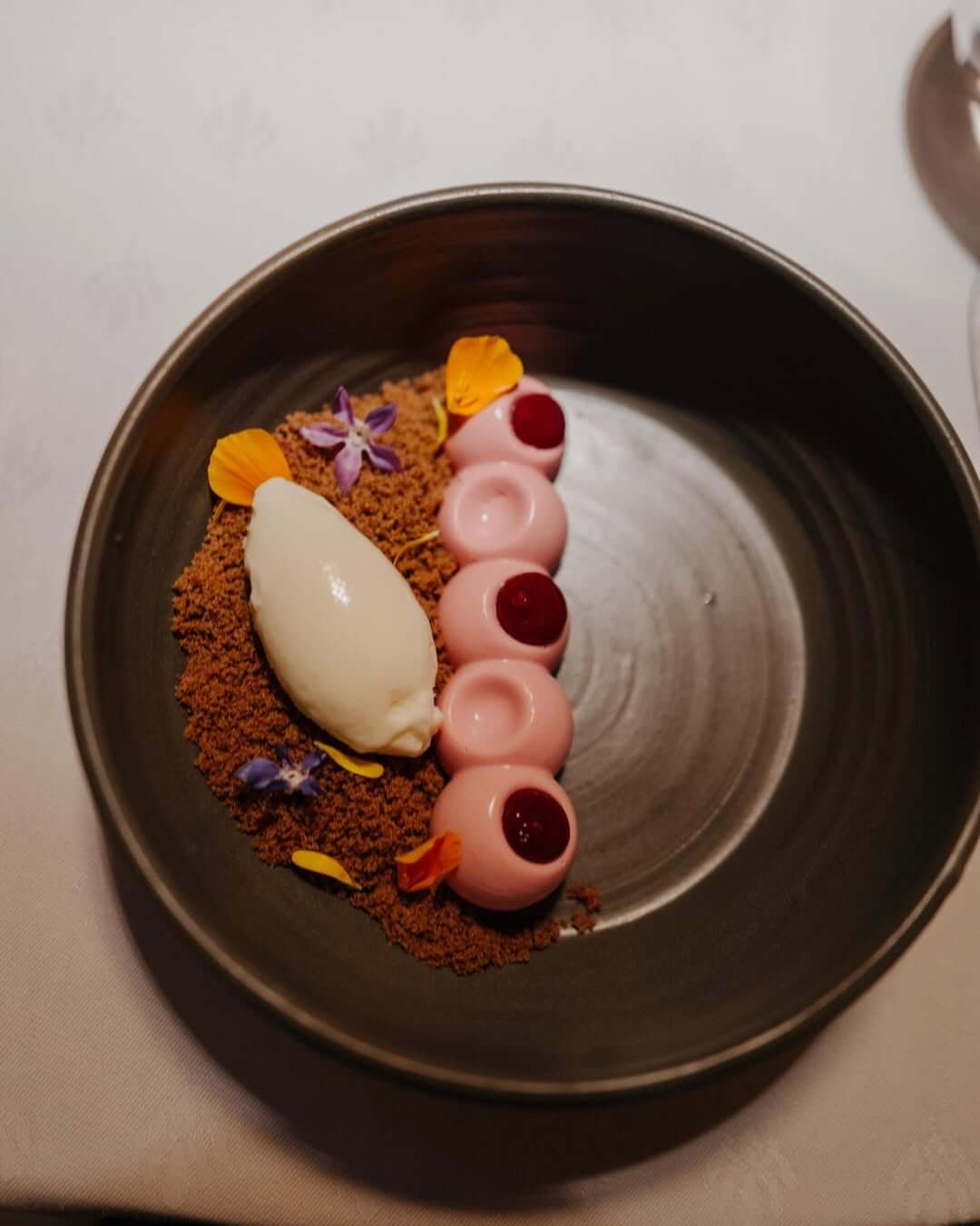 Modern plated dessert with pink mousse domes, berry centers, chocolate crumble, sorbet, and edible flowers in a dark bowl.
