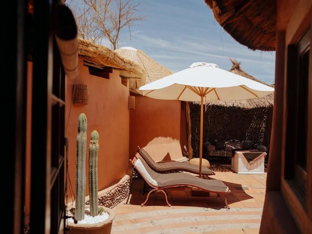 Sunlit adobe courtyard with lounge chairs, white umbrella, cacti, and thatched roofs creating a serene desert retreat.