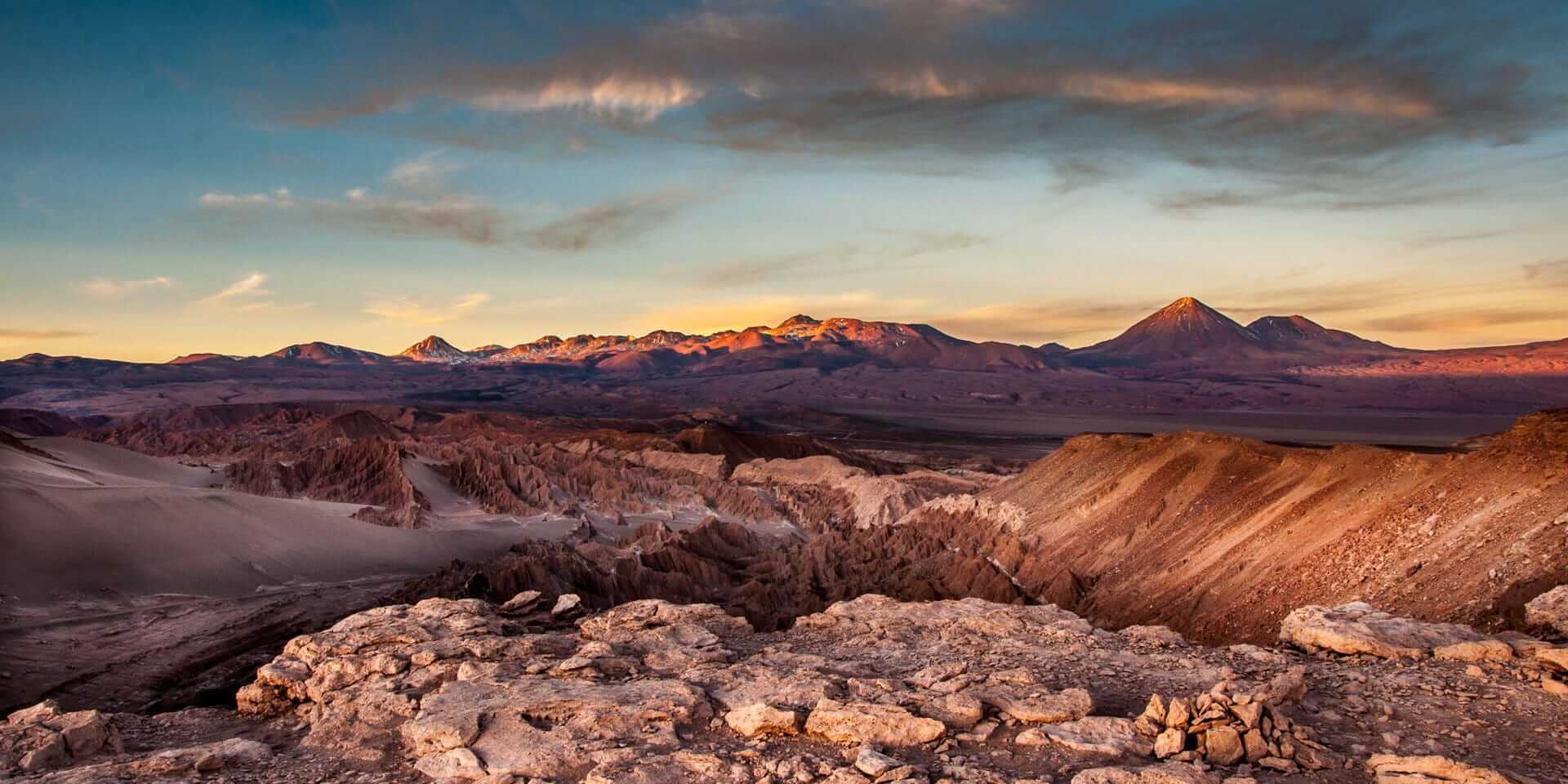 Panoramic desert landscape at sunset with rugged canyons, salt formations, and distant mountains glowing in warm light.