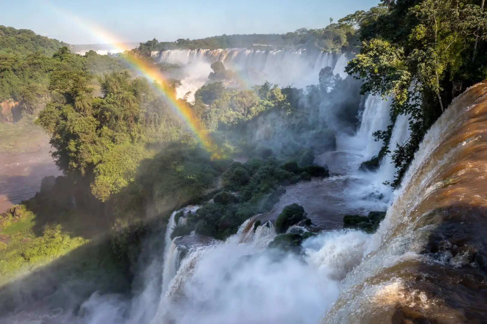 Powerful waterfalls cascading through lush rainforest with mist rising and a rainbow arching across the scene.