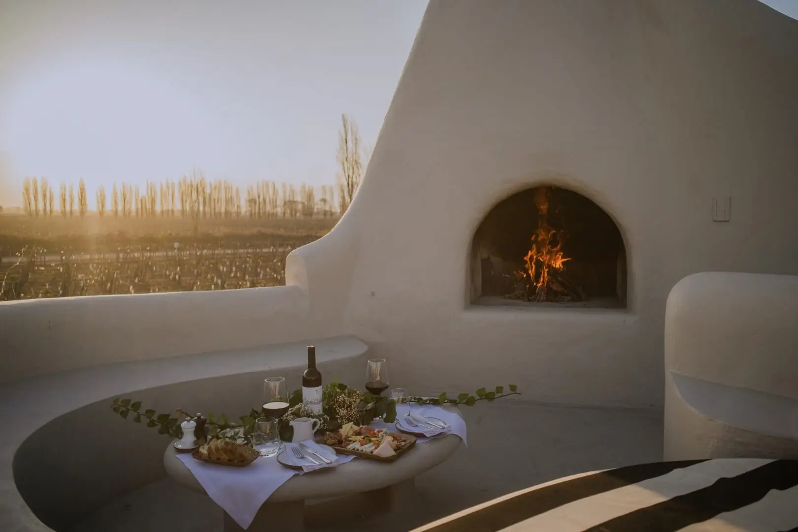 Cozy outdoor table set beside a white adobe fireplace with a burning fire, overlooking vineyards at sunset.