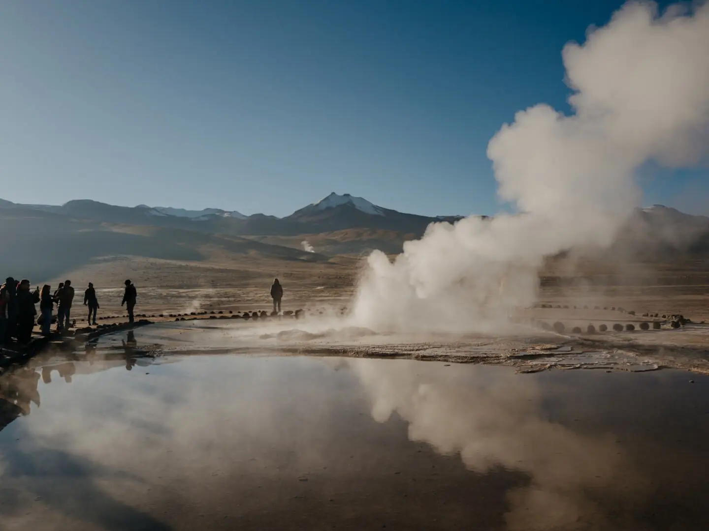 Steam rises from a geothermal pool as visitors stand nearby, reflected in still water beneath distant andean peaks.
