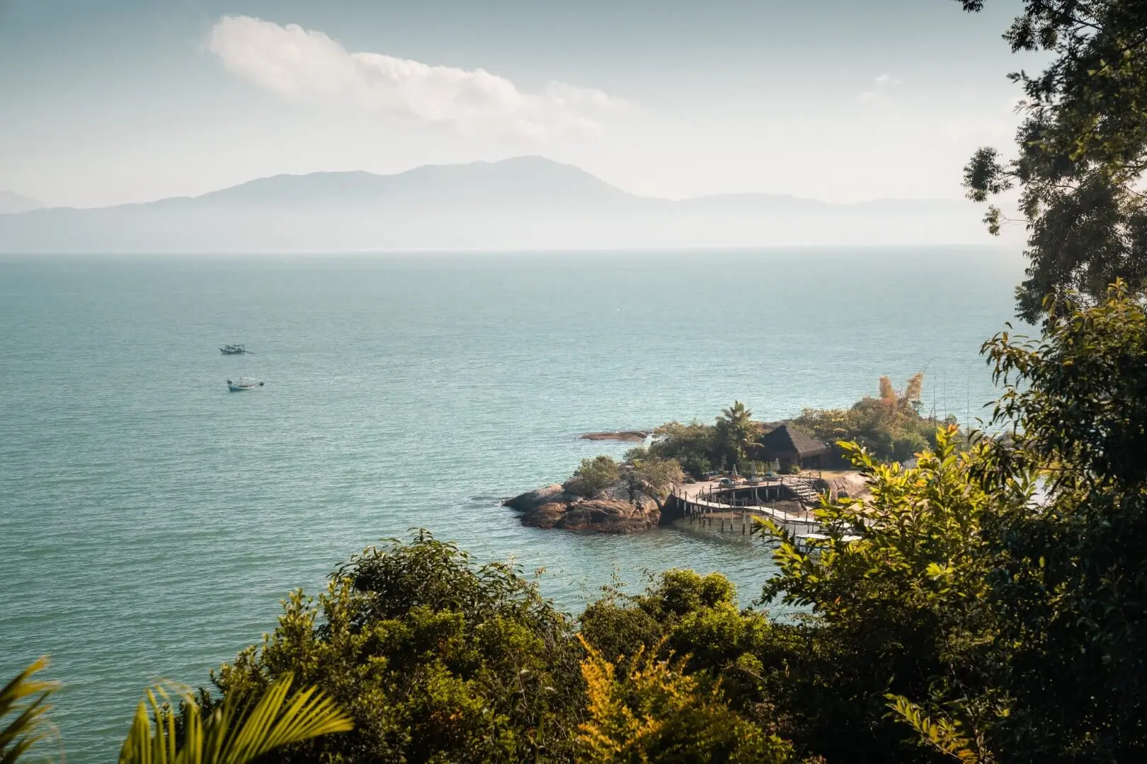 Coastal overlook with calm blue sea, small rocky peninsula and dock, boats offshore, distant mountains, and lush green foliage.