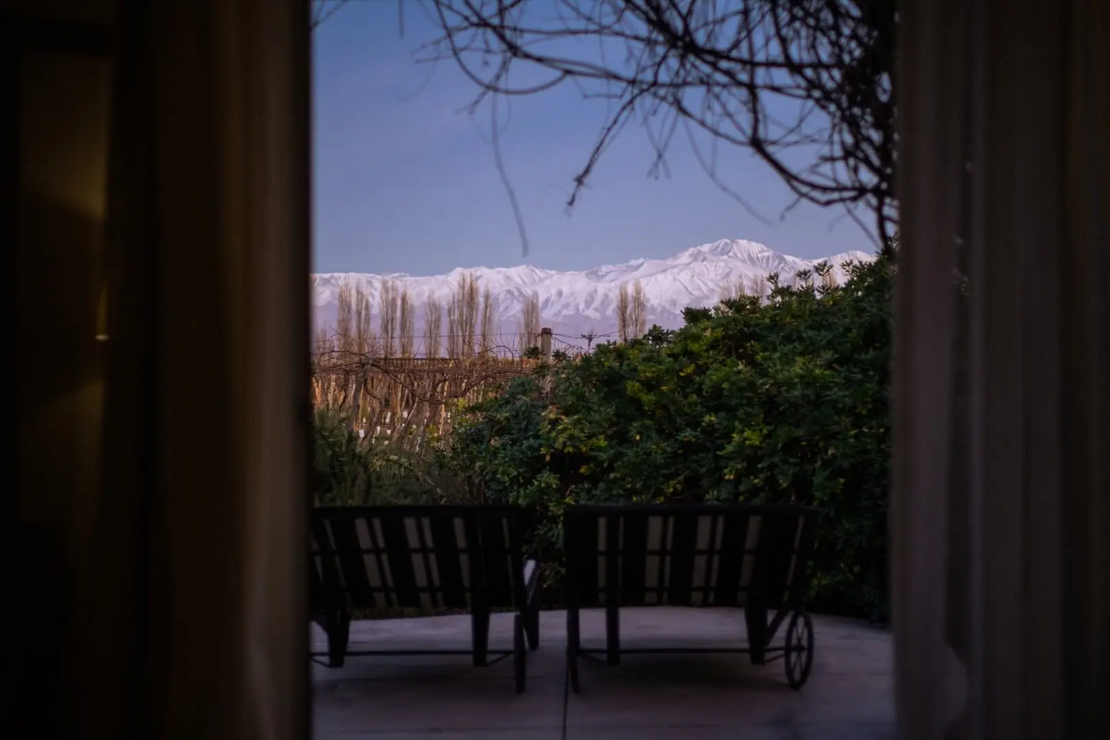 Snow-capped mountains viewed from a shaded patio with lounge chairs, framed by garden greenery at dusk.