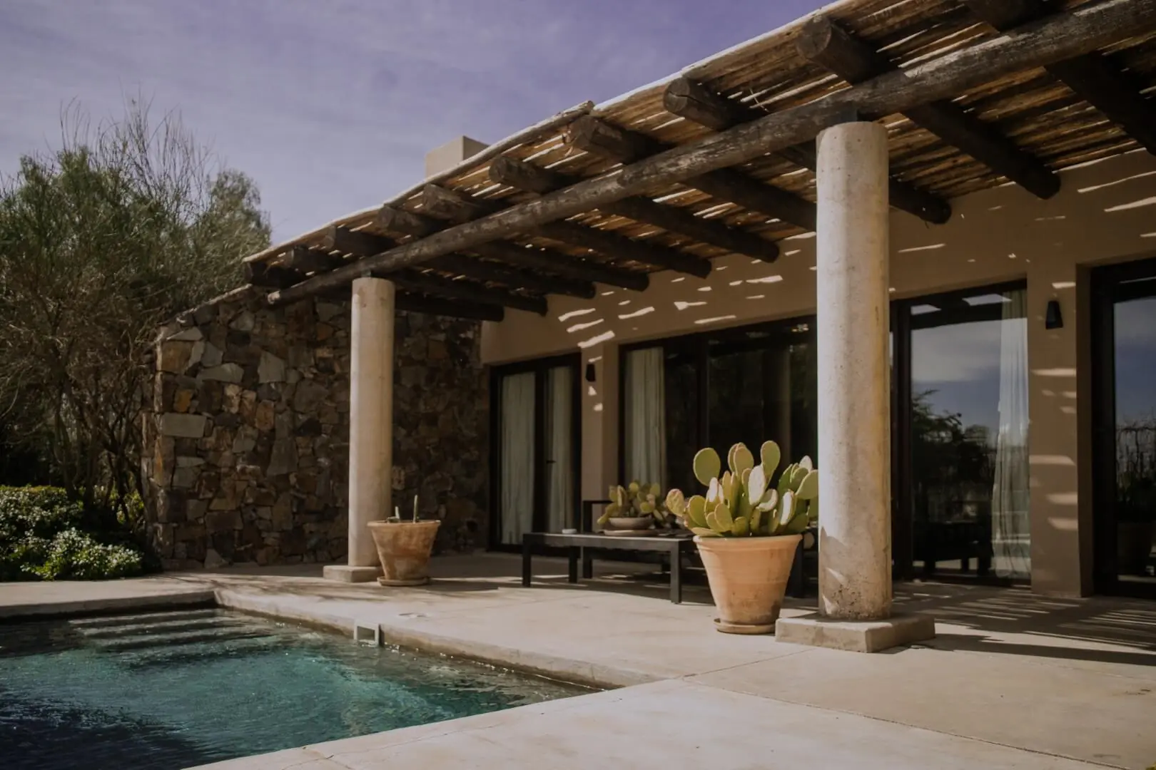 Stone-and-adobe villa with shaded terrace, poolside seating, and cactus planters under a wooden pergola.