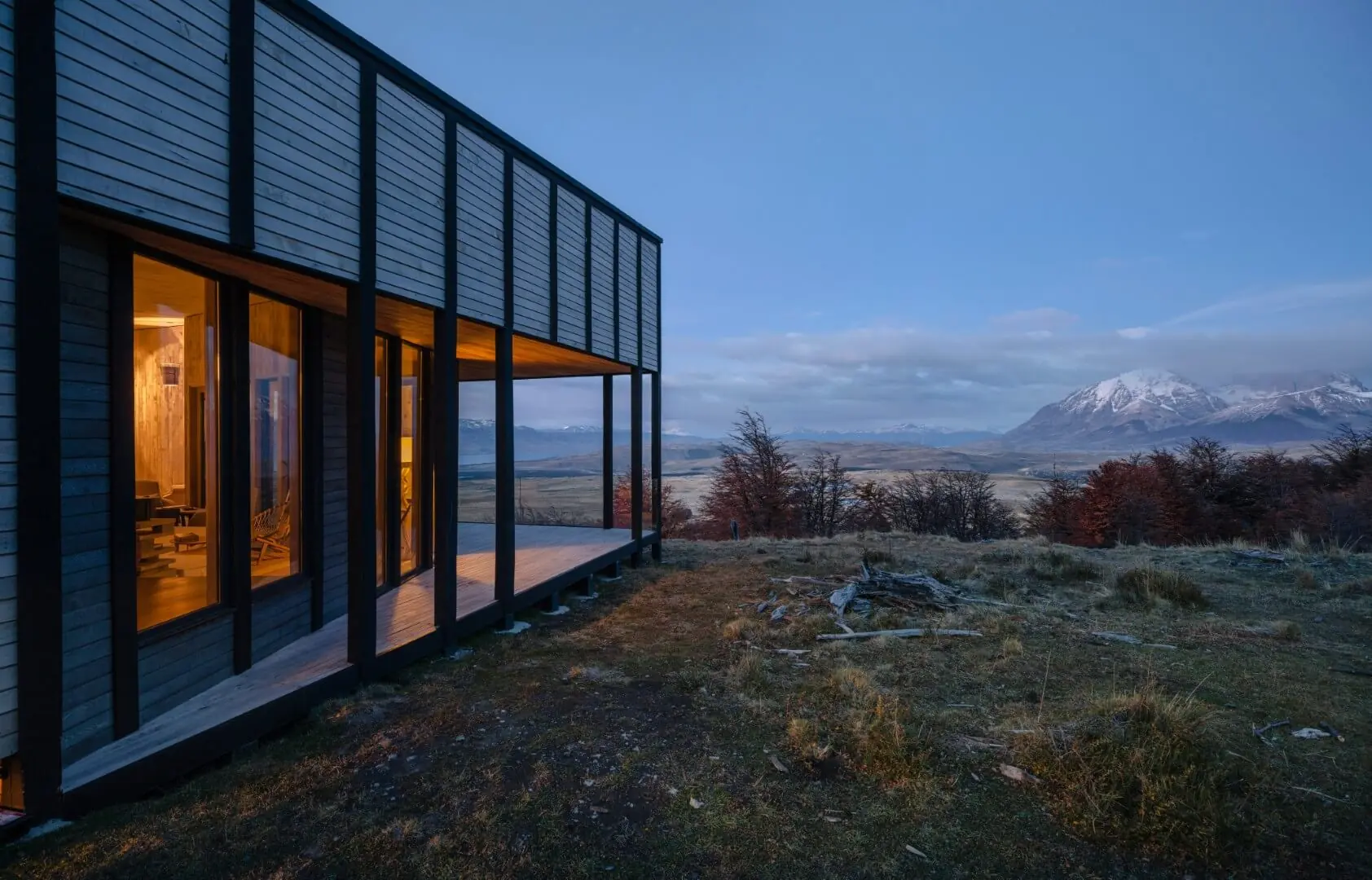 Modern cabin with large windows overlooking a vast landscape and snow-capped mountains at dusk.