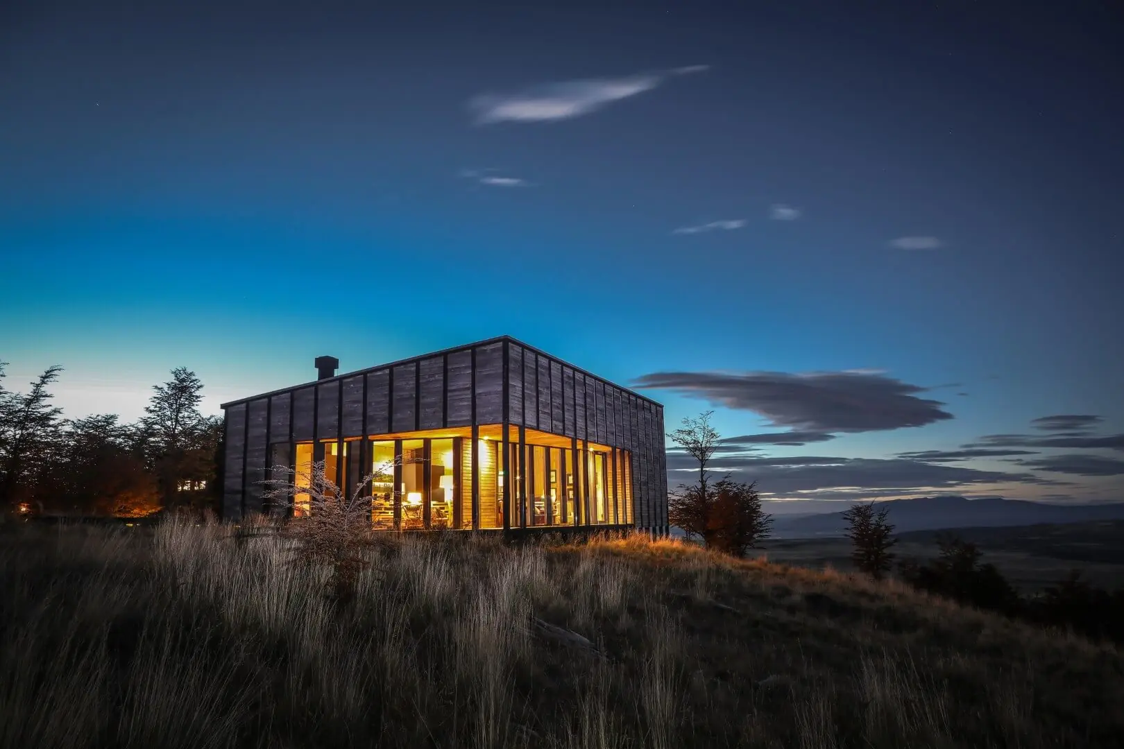 Modern glass cabin glowing at dusk, set on a grassy hillside beneath a deep blue evening sky.