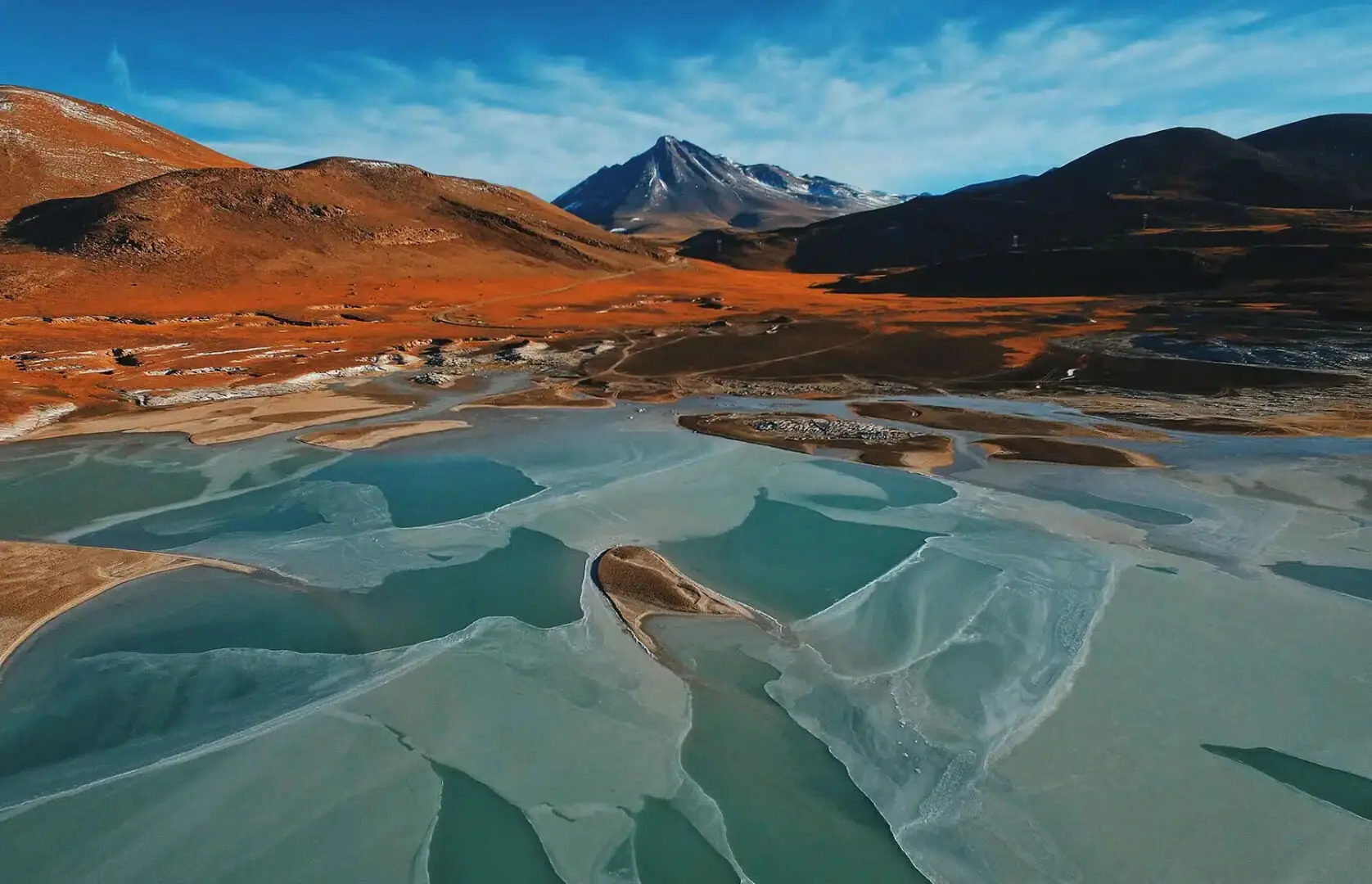 Aerial view of turquoise river channels winding through a red desert valley with mountains under a clear blue sky.