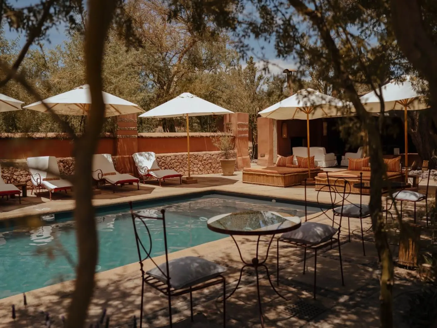 Sunlit courtyard pool with lounge chairs, white umbrellas, and cozy seating surrounded by trees and earth-toned walls.