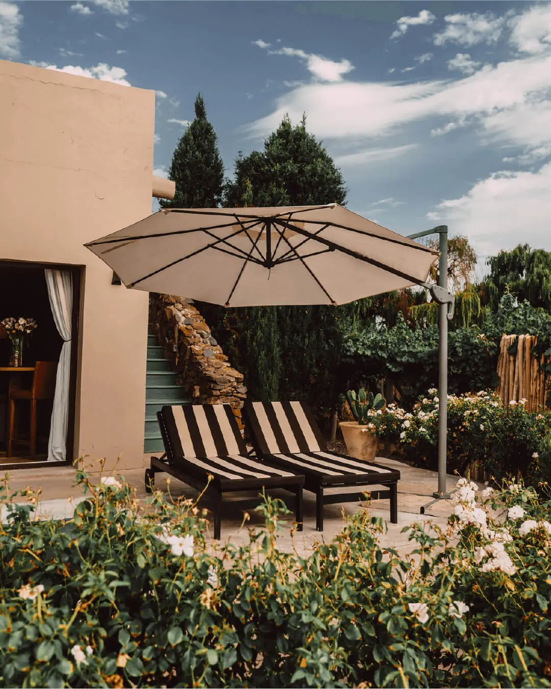 Two striped lounge chairs beneath a patio umbrella outside a villa, surrounded by garden greenery and flowers
