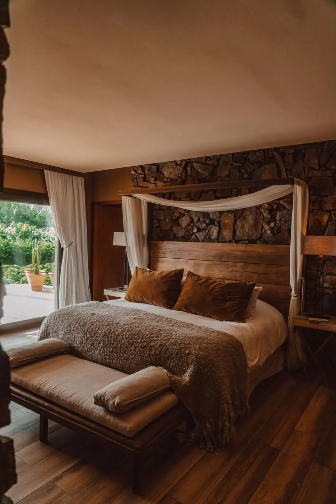 Cozy rustic bedroom with wooden bed, stone accent wall, soft canopy drapes, and warm natural light.