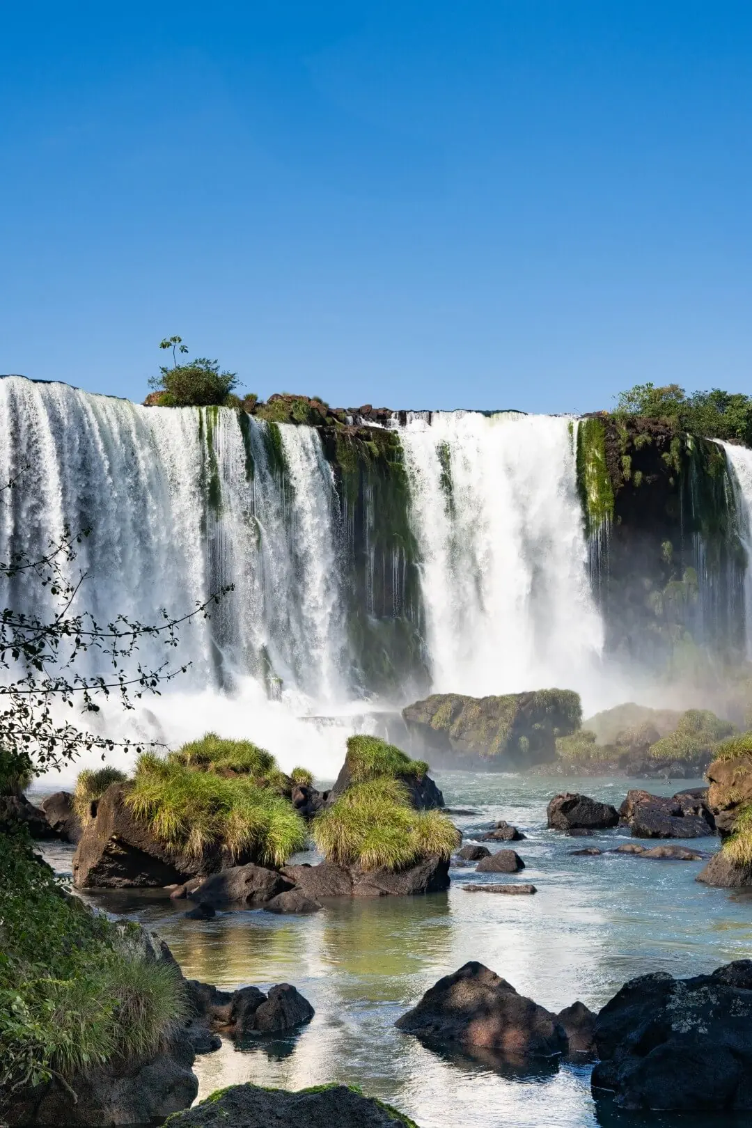 Wide Waterfall Cascading Over Cliffs Into Rocky River With Lush Greenery Under Clear Blue Sky.