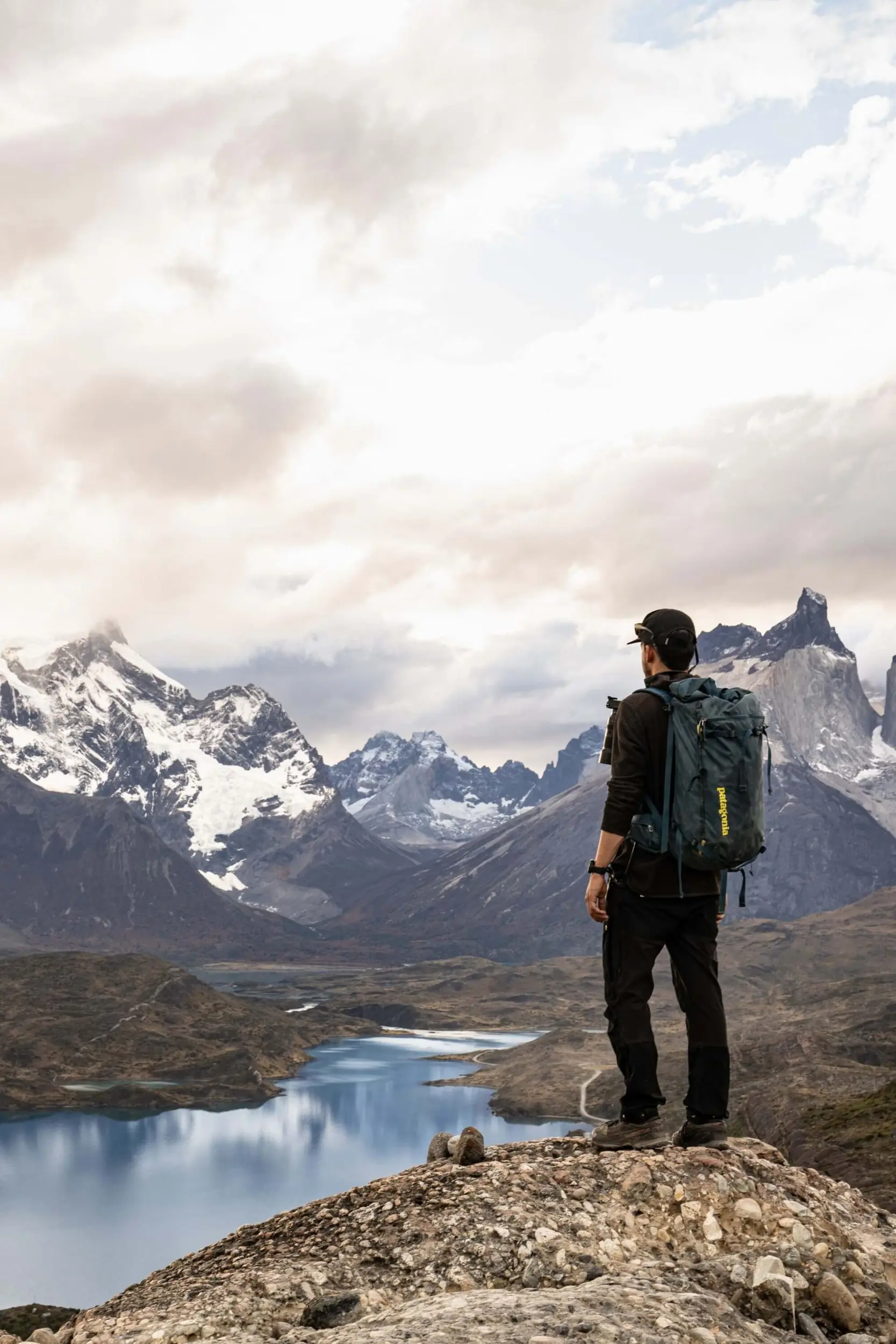 Hiker with backpack standing on a rocky ridge overlooking a blue lake and snow-capped mountains.