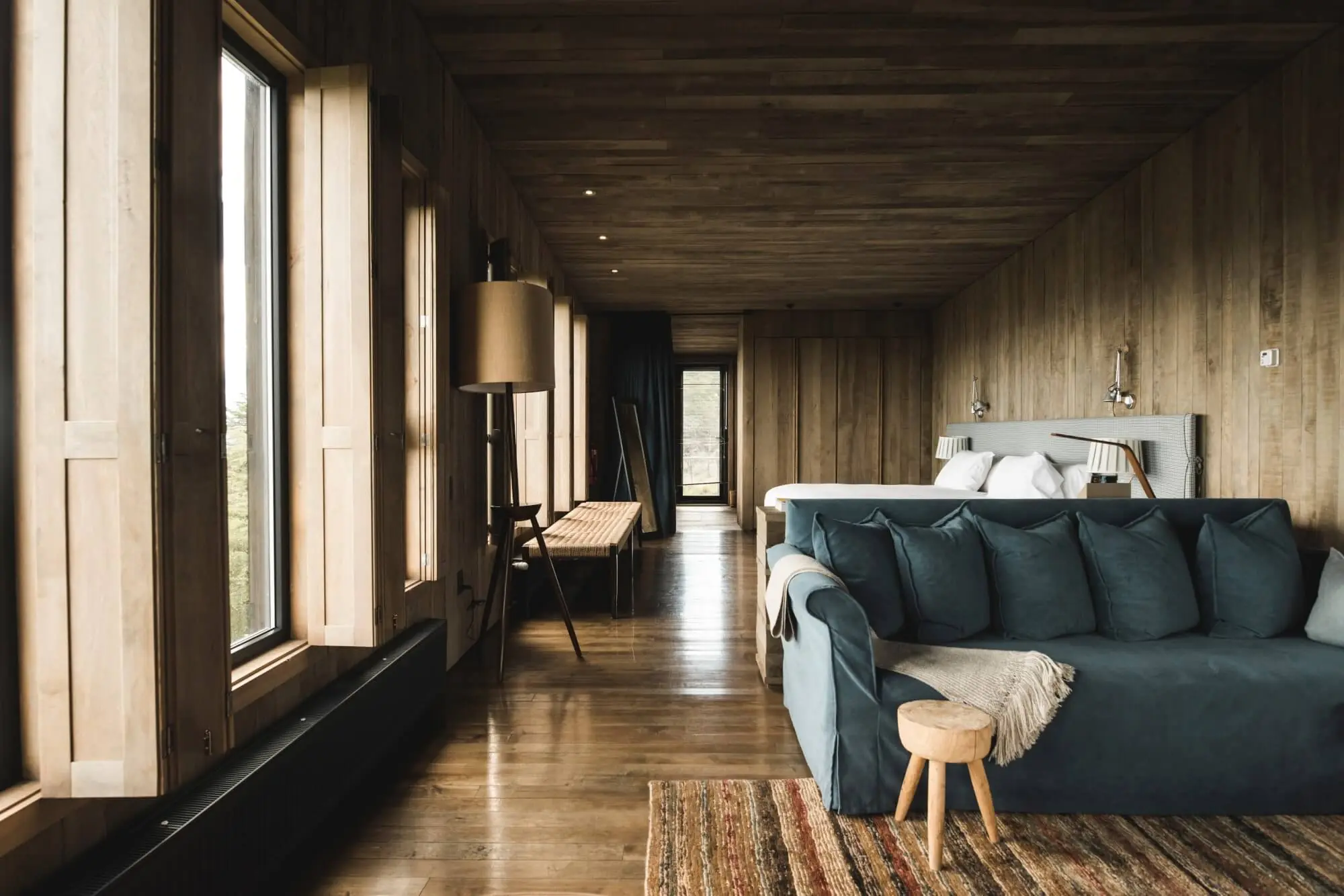 Modern rustic bedroom with wood-paneled walls, large windows, blue sofa, hardwood floors, and a bed at the far end.