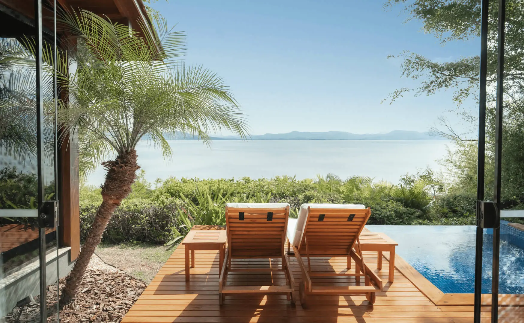 Two wooden lounge chairs by a private pool overlooking a calm lake, framed by palm trees and clear blue sky.