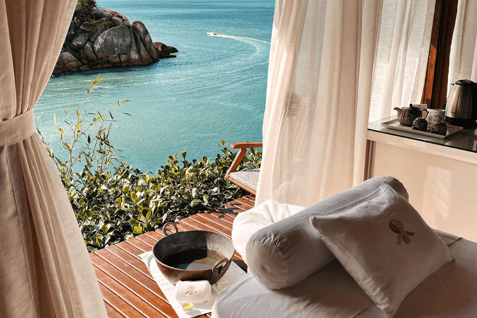 Ocean-view spa setup with massage bed, sheer curtains, wooden deck, and turquoise water with a rocky islet outside