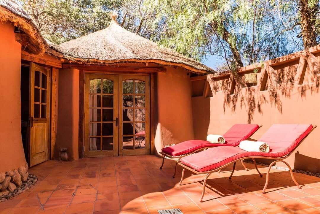 Private sun patio with two red loungers, terracotta tiles, and a thatched-roof adobe suite entrance.
