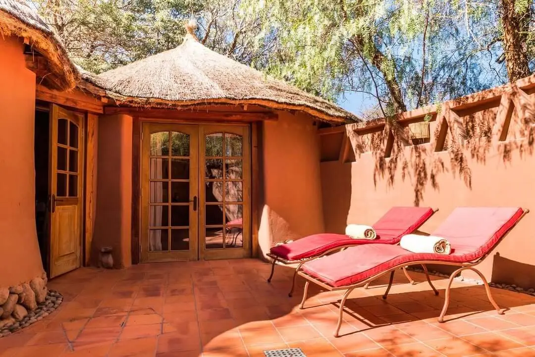 Private sun patio with two red loungers, terracotta tiles, and a thatched-roof adobe suite entrance.