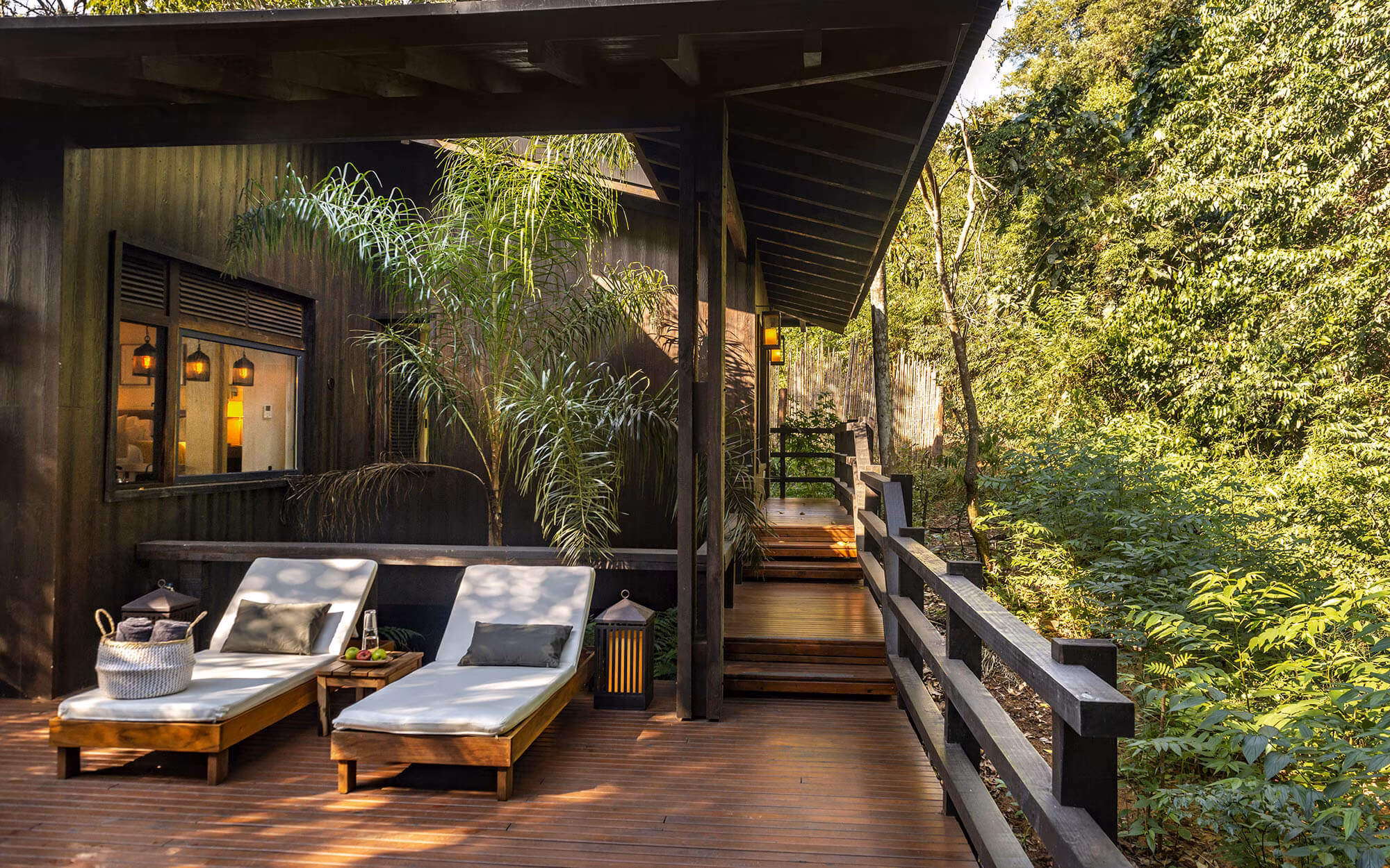 Wooden Forest Cabin Deck With Two Lounge Chairs, Soft Cushions, And A Walkway Surrounded By Lush Green Foliage.