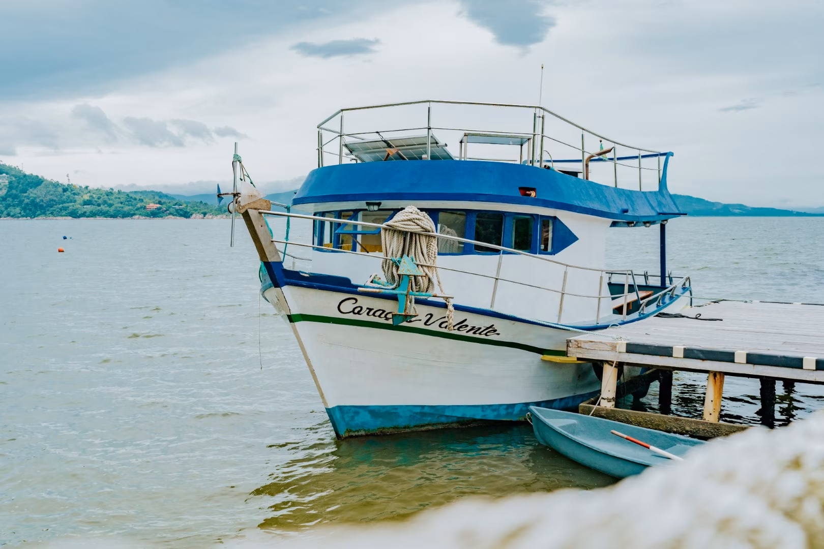 Blue and white boat docked at a wooden pier on calm water, with hills and cloudy sky in the background.