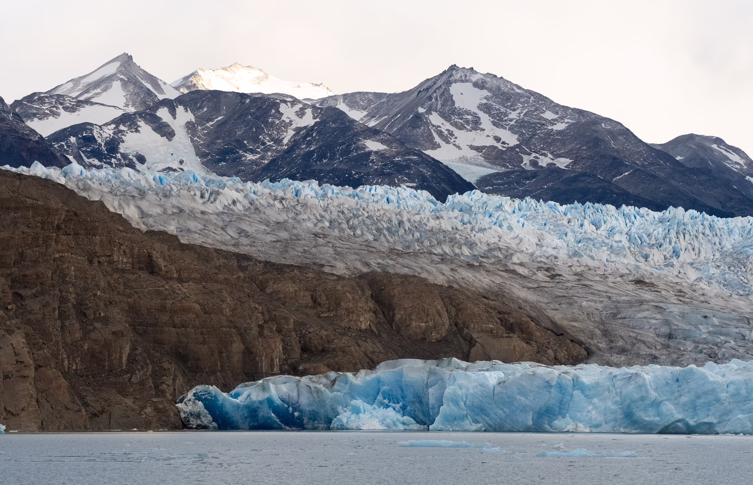Blue glacier meets rocky cliffs and snow-capped mountains beside a calm lake.