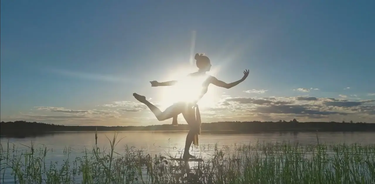 Silhouetted dancer balancing on lakeshore at sunset with sunburst behind against calm water.