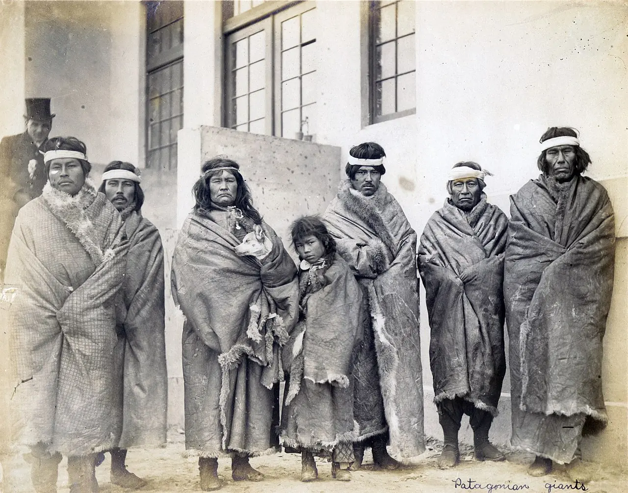 Historic black-and-white photo of Indigenous Patagonian group wrapped in blankets outside building.