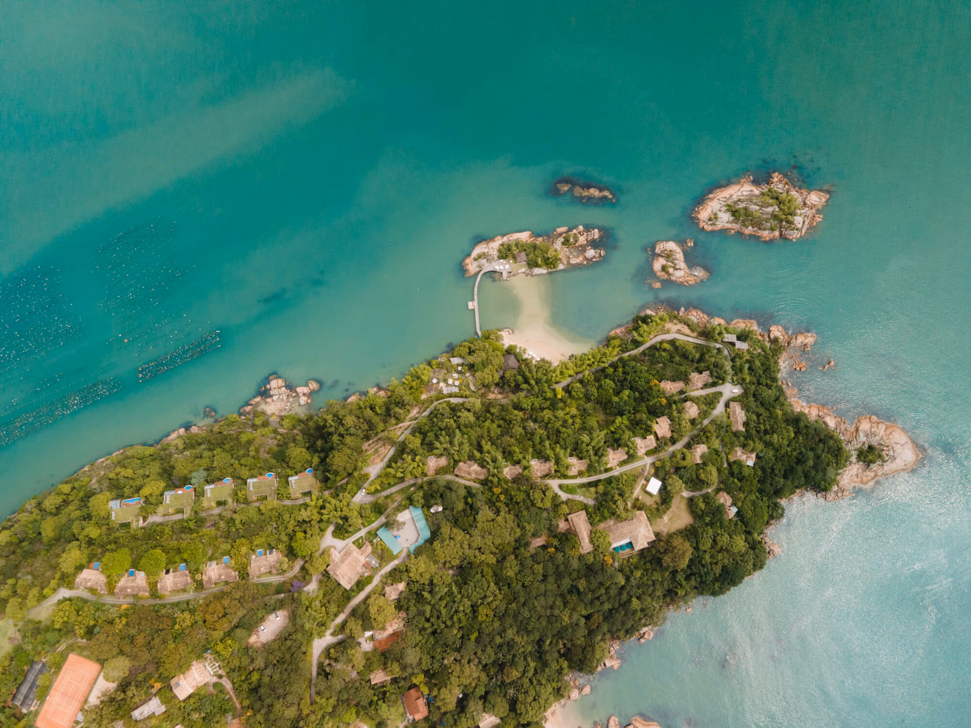 Aerial view of a lush island resort with villas, winding paths, and turquoise bays along a rocky coastline.