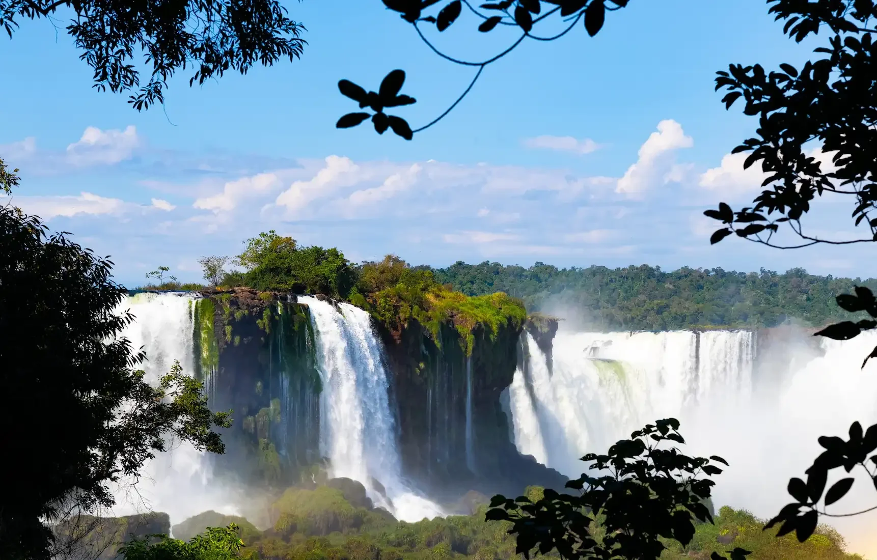 Wide waterfall cascading over lush cliffs, framed by leafy trees under a bright blue sky.